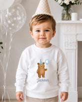 A young boy wearing a white sweatshirt with a teddy bear and a birthday hat, smiling at the camera.