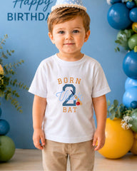 A young boy wearing a white t-shirt with the text "BORN 2 BAT" stands in front of a blue background with balloons and greenery.