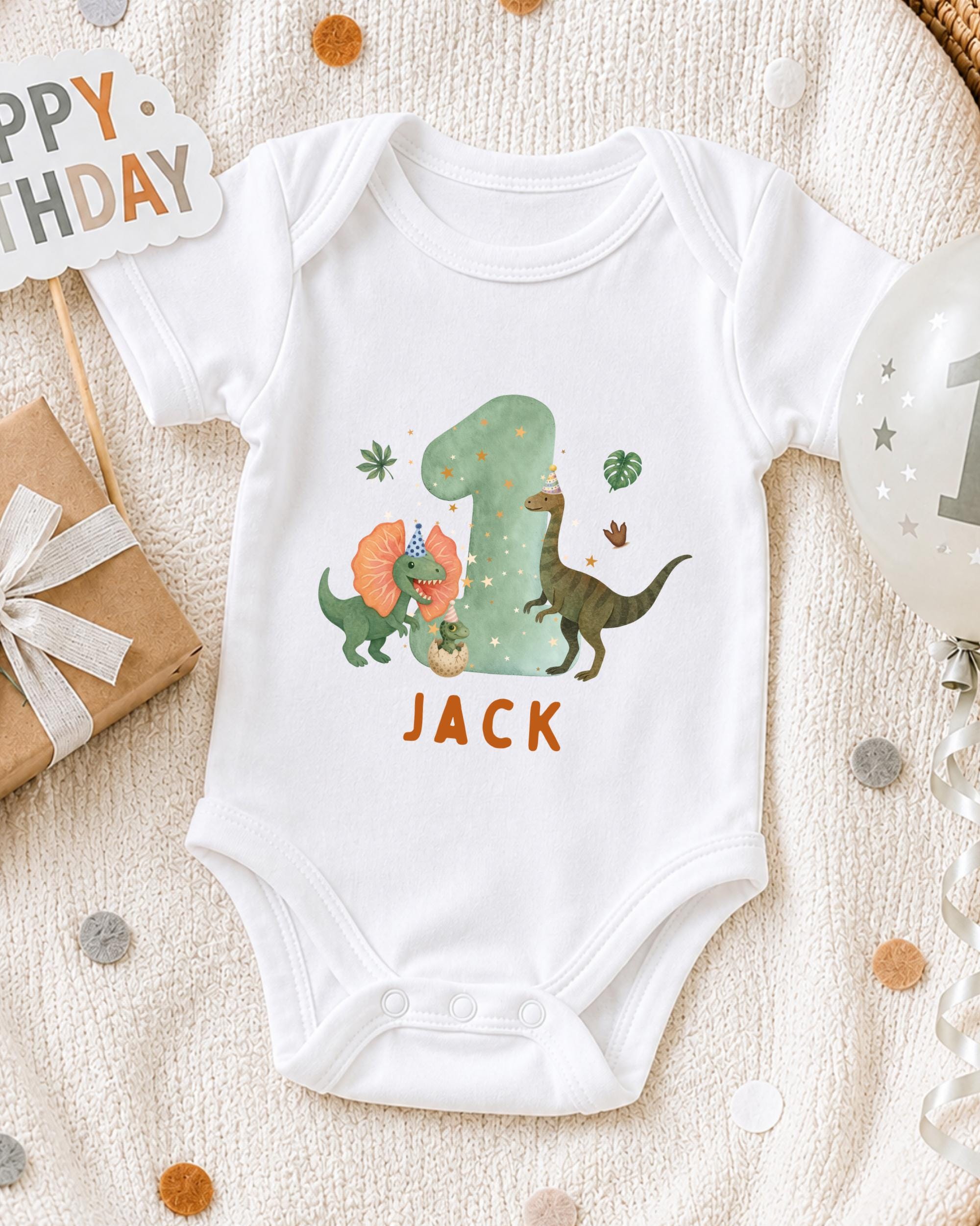 A white onesie with a dinosaur design and the name "Jack" printed on it, along with a birthday sign and a balloon, suggesting a celebration for a baby's first birthday.