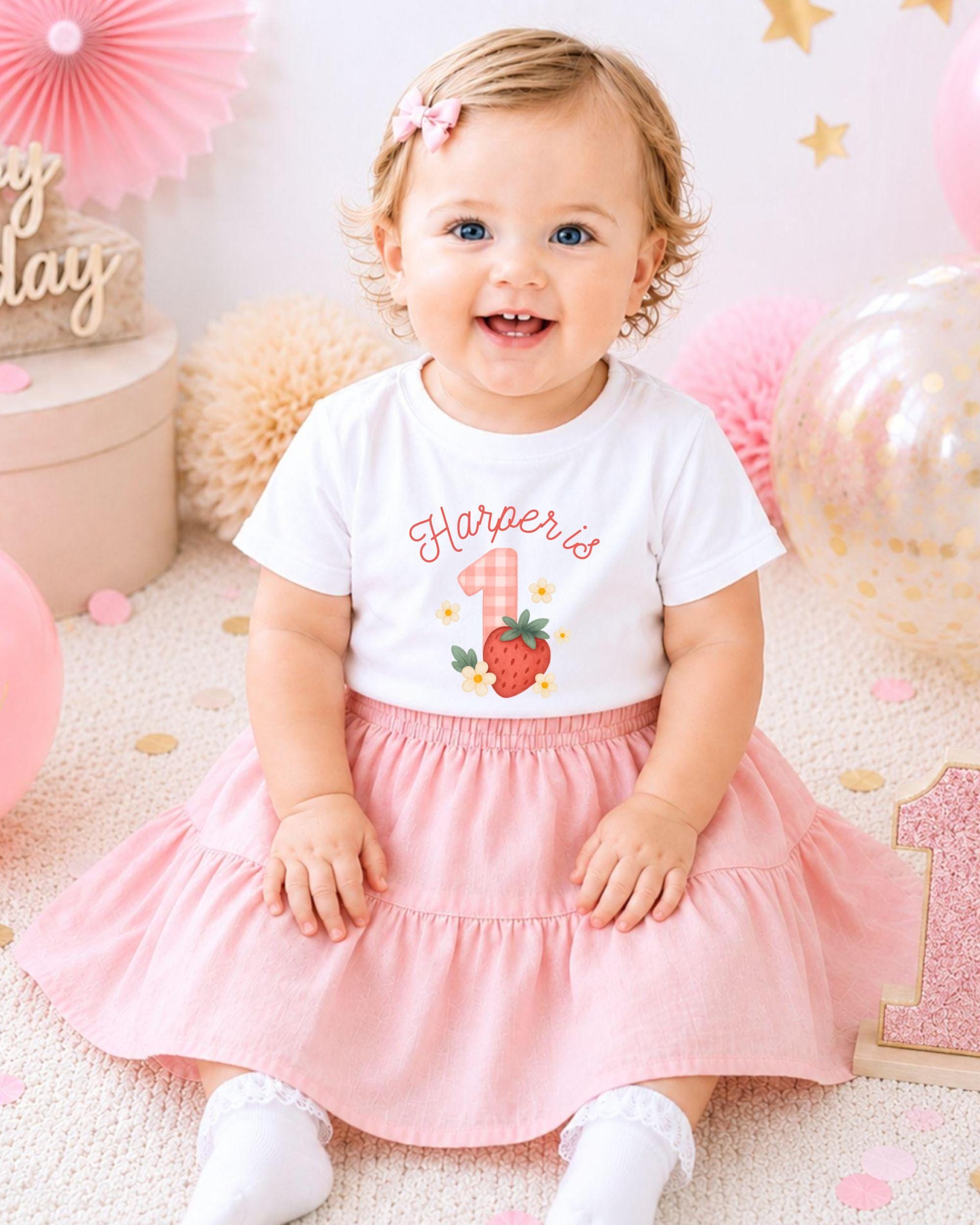 A smiling baby girl wearing a white t-shirt with a strawberry design and a pink skirt, sitting on a pink and white background with balloons and confetti.