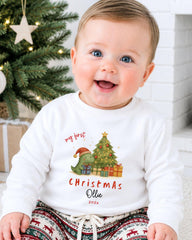 A smiling baby wearing a white Christmas-themed sweatshirt with a dinosaur and presents, sitting in front of a Christmas tree.