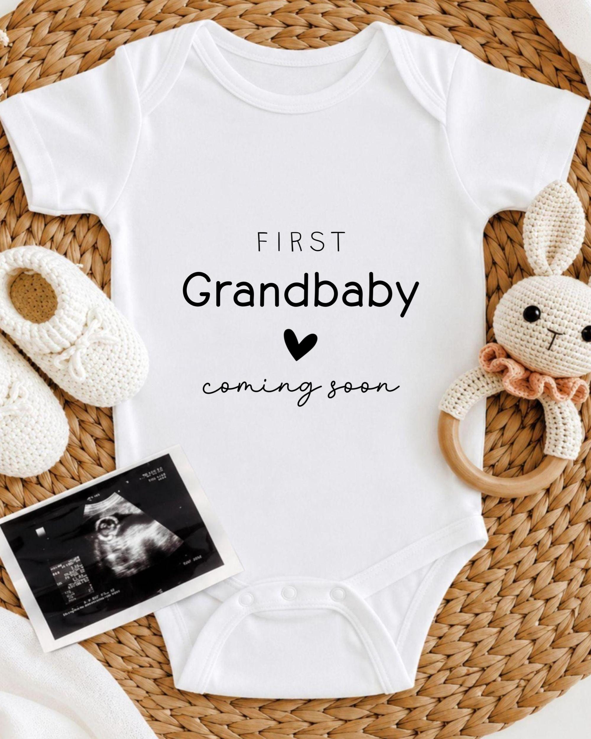A white baby onesie with the text "FIRST Grandbaby coming soon" printed on it, along with a crocheted rabbit toy and a black and white photograph of an infant's head.