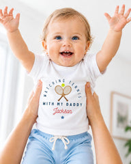 A smiling baby wearing a white shirt with a tennis racket and the text "Watching Tennis with my Daddy Jackson" is being held up by someone's arms.