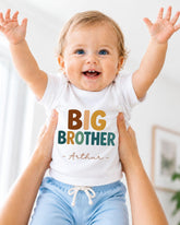 A smiling baby wearing a white shirt with the text "BIG BROTHER - ARTHUR" is being held up by someone's arms.
