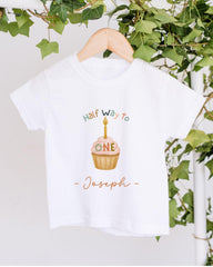 A white t-shirt with a graphic design featuring a cupcake with a candle on it, along with the text "Half Way to Joseph" printed on it.