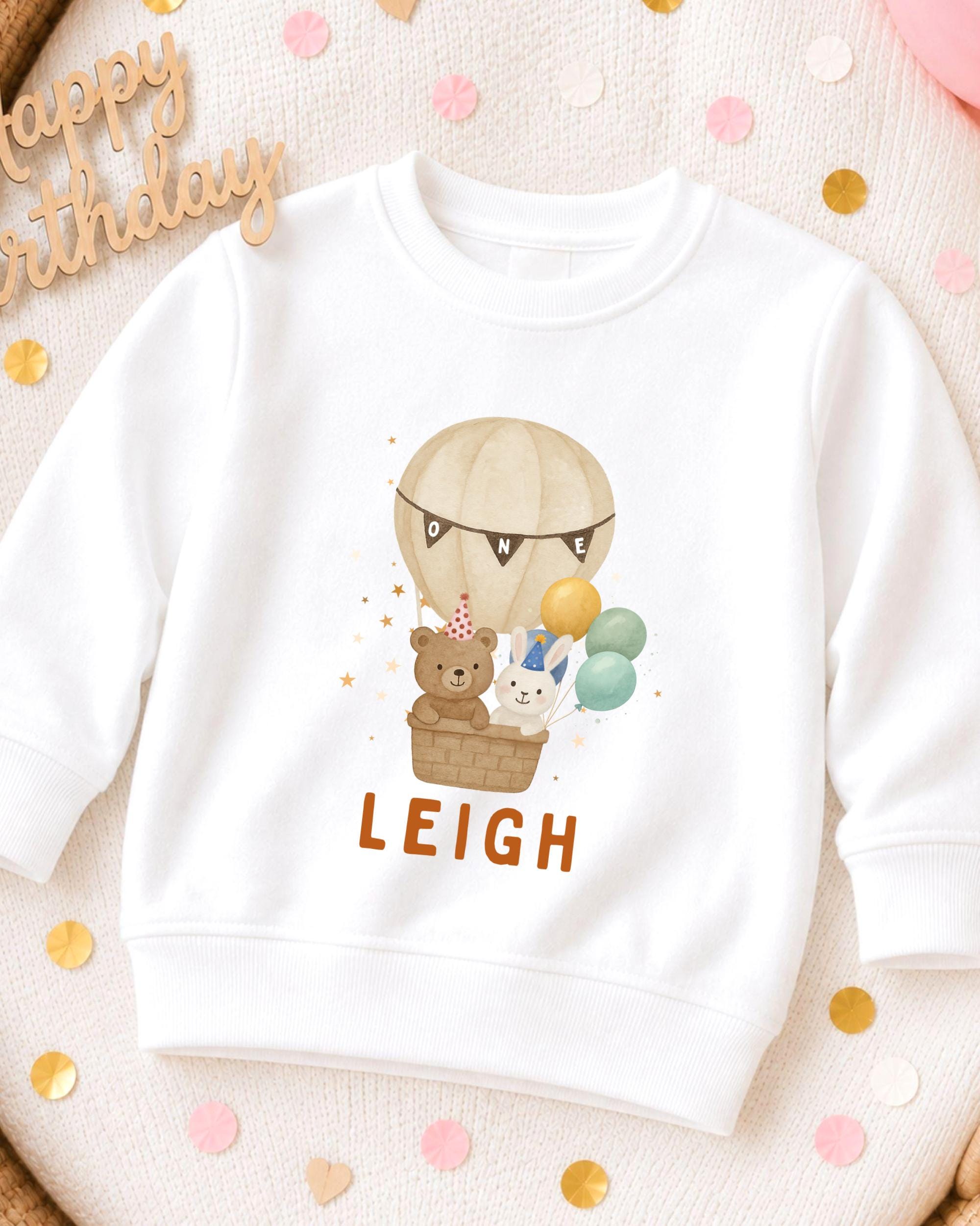 A white sweatshirt with a cartoon image of a hot air balloon carrying a teddy bear and a rabbit, along with the name "LEIGH" printed on it.