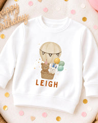 A white sweatshirt with a cartoon image of a hot air balloon carrying a teddy bear and a rabbit, along with the name "LEIGH" printed on it.