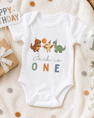 A white onesie with a dinosaur and a party hat printed on it, along with a "Jack is one" message, is displayed on a bed with a "Happy Birthday" sign and a balloon.