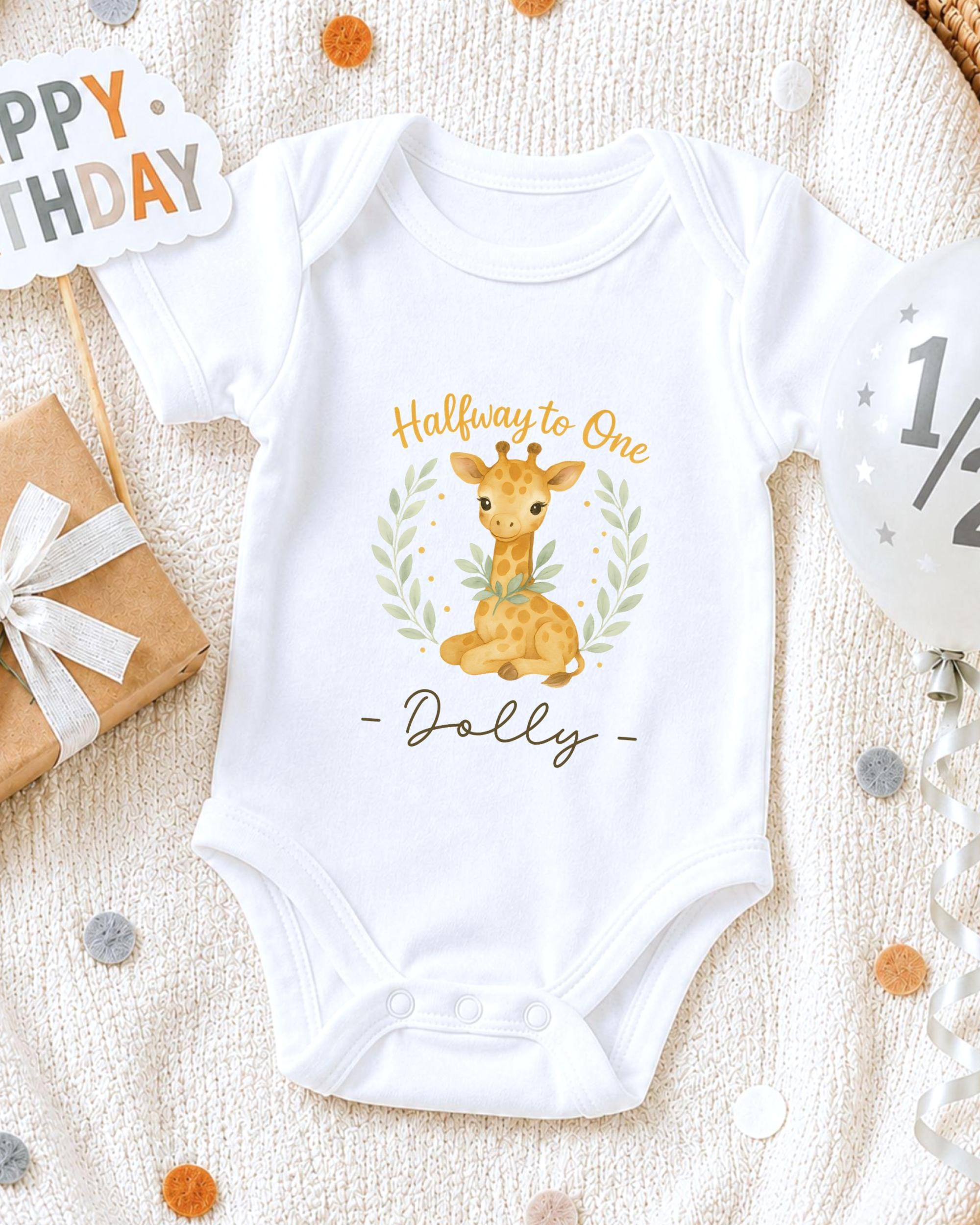A white onesie with a giraffe design and the text "Halfway to One Dolly" printed on it, along with a "Happy Birthday" sign and confetti.
