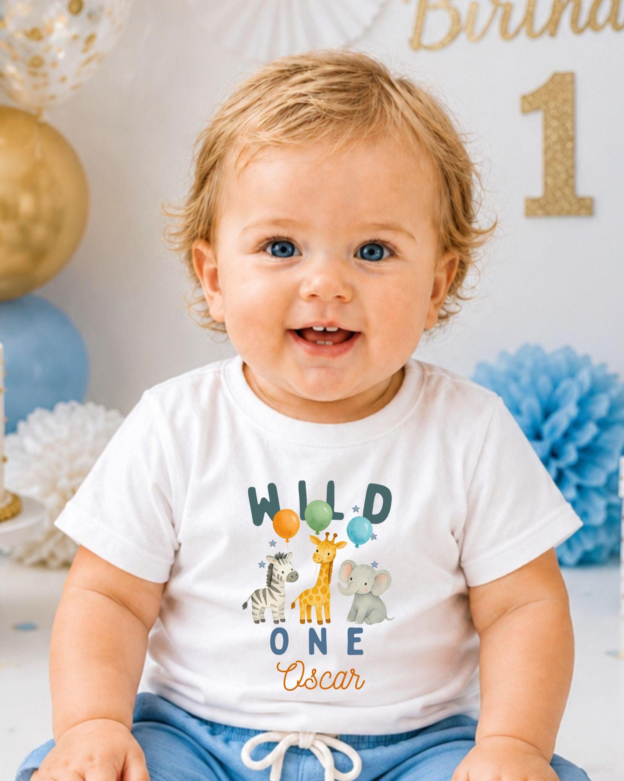 A smiling baby boy wearing a white t-shirt with a cartoon animal design and the text "Wild One Oscar" is sitting in front of a birthday party decoration.