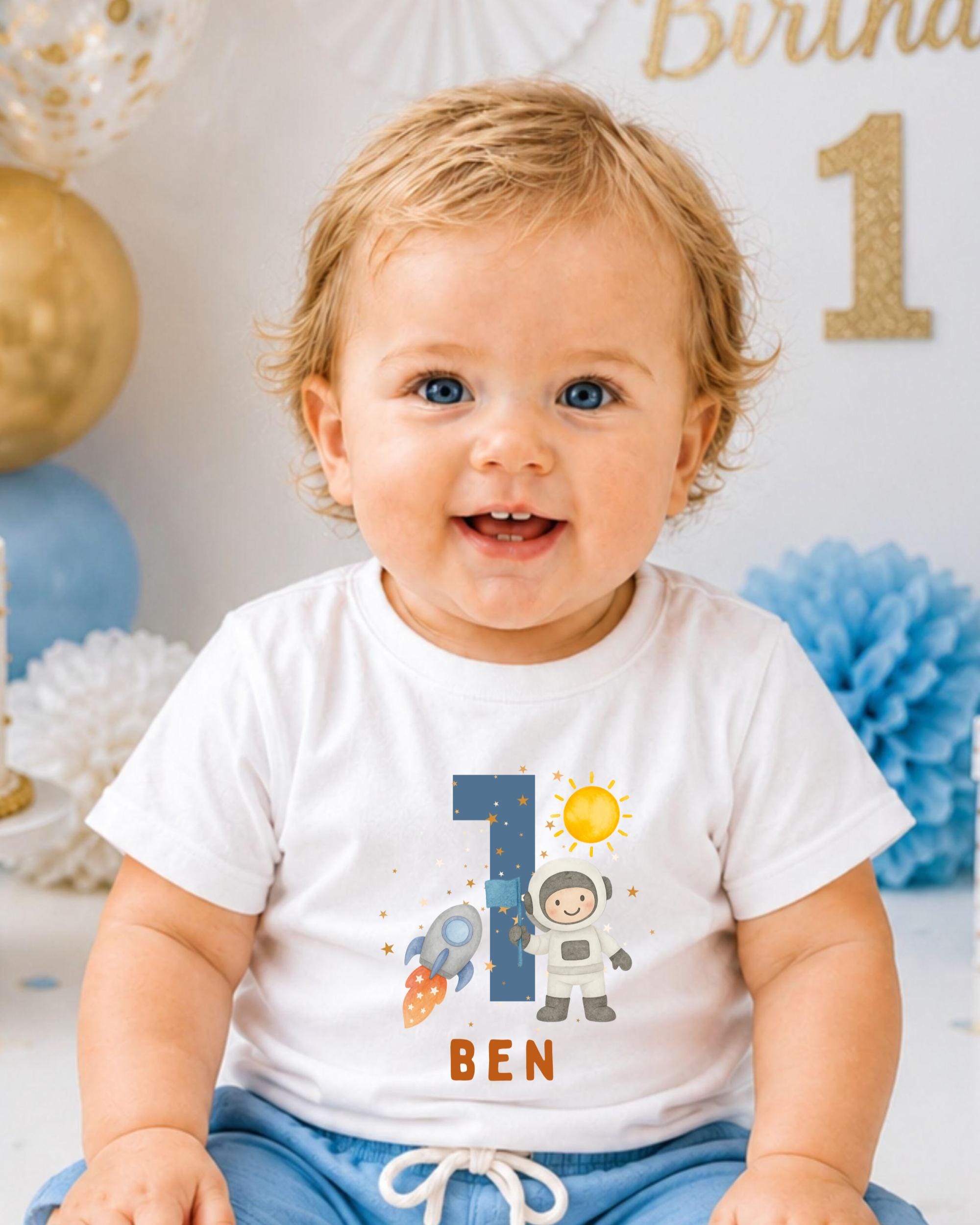A smiling baby boy wearing a white t-shirt with a space-themed design and the number 1, sitting in front of a birthday party backdrop.