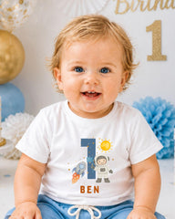 A smiling baby boy wearing a white t-shirt with a space-themed design and the number 1, sitting in front of a birthday party backdrop.