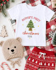 A white t-shirt with a Christmas tree design, a beige knit hat with a bear face, and red and white patterned shorts are arranged on a plaid blanket, along with Christmas decorations and ornaments.