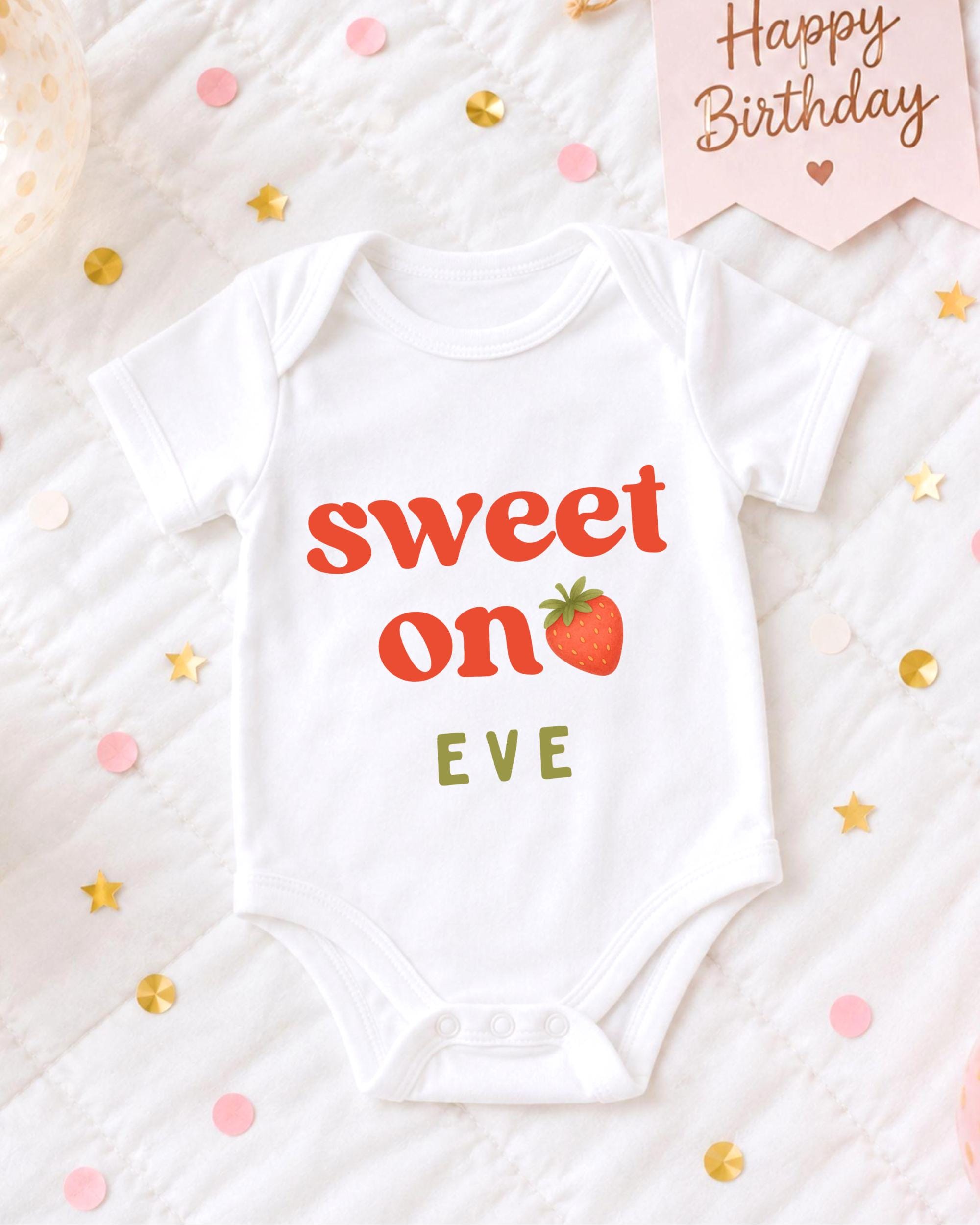 A white baby onesie with the text "Sweet on EVE" printed on it, placed on a bed with confetti and a birthday card.