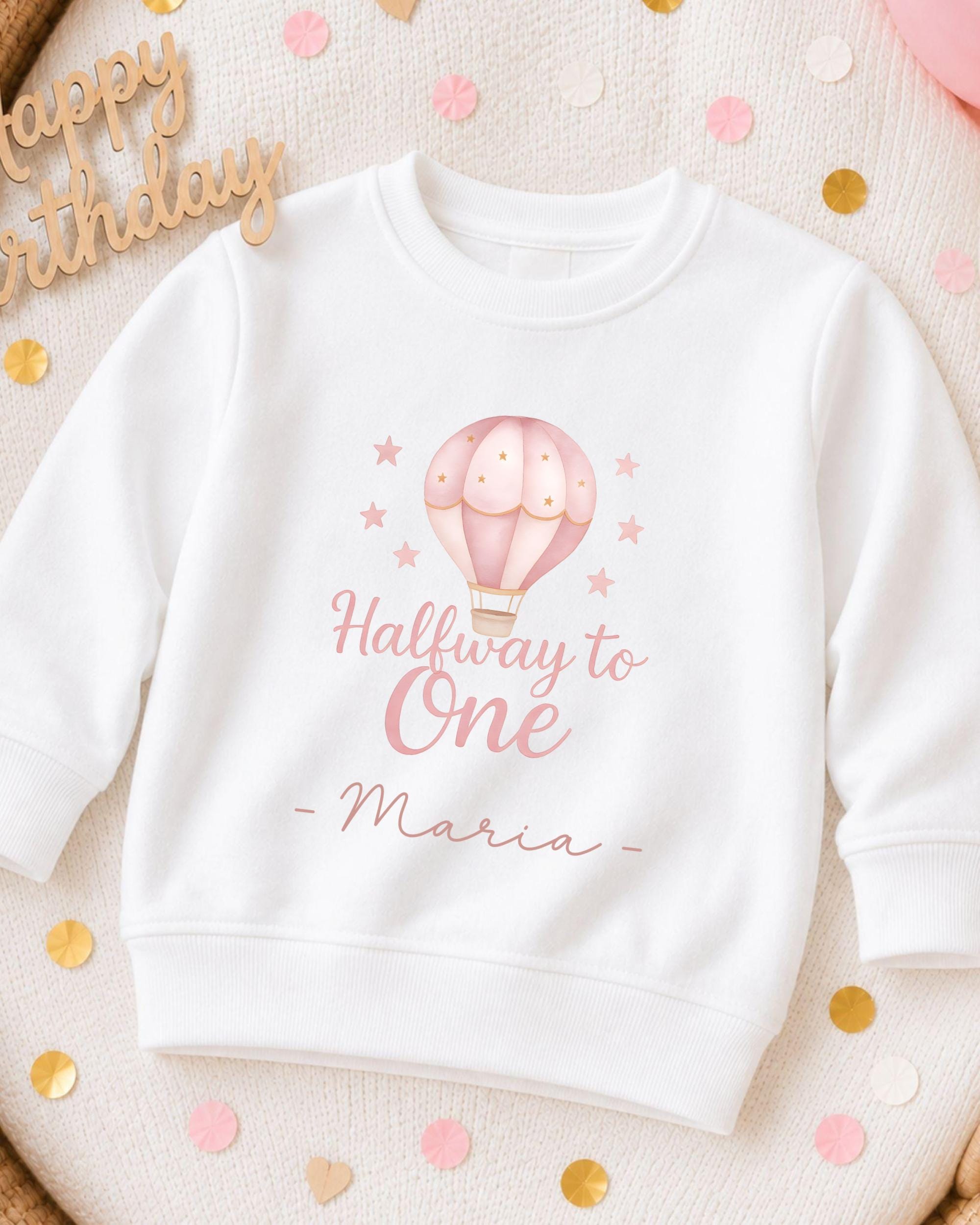 A white sweatshirt with a pink hot air balloon design and the text "Halfway to One - Maria" printed on it, surrounded by confetti.