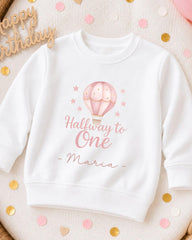 A white sweatshirt with a pink hot air balloon design and the text "Halfway to One - Maria" printed on it, surrounded by confetti.