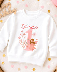 A white sweatshirt with a pink fairy and the number 1 printed on it, surrounded by confetti.