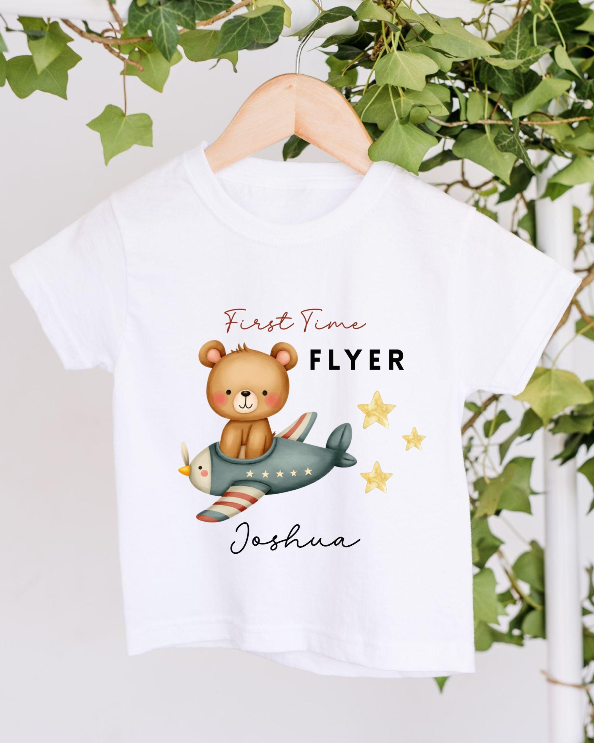 A white t-shirt with a cartoon image of a bear riding a plane and stars, along with the text "First Time Flyer Joshua" printed on it.