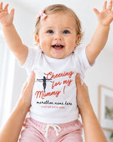 A smiling baby girl wearing a white t-shirt with a cheerleading design and pink shorts, being held up by someone's arms.