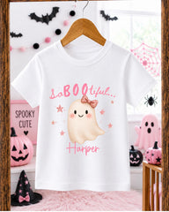 A white t-shirt with a pink ghost and the text "SaBOE LITERAL Harper" printed on it, hanging on a wooden hanger. The t-shirt is displayed in a room decorated for Halloween, with various Halloween-themed decorations and items visible in the background.