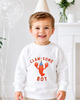 A young boy wearing a white sweatshirt with a cartoon lobster and the text "CLAW SOME BOY" is smiling at the camera.