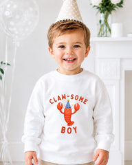 A young boy wearing a white sweatshirt with a cartoon lobster and the text "CLAW SOME BOY" is smiling at the camera.