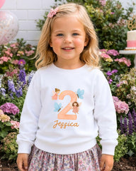A young girl wearing a white sweater with a number "2" and a fairy design, standing in front of a garden with colorful flowers.