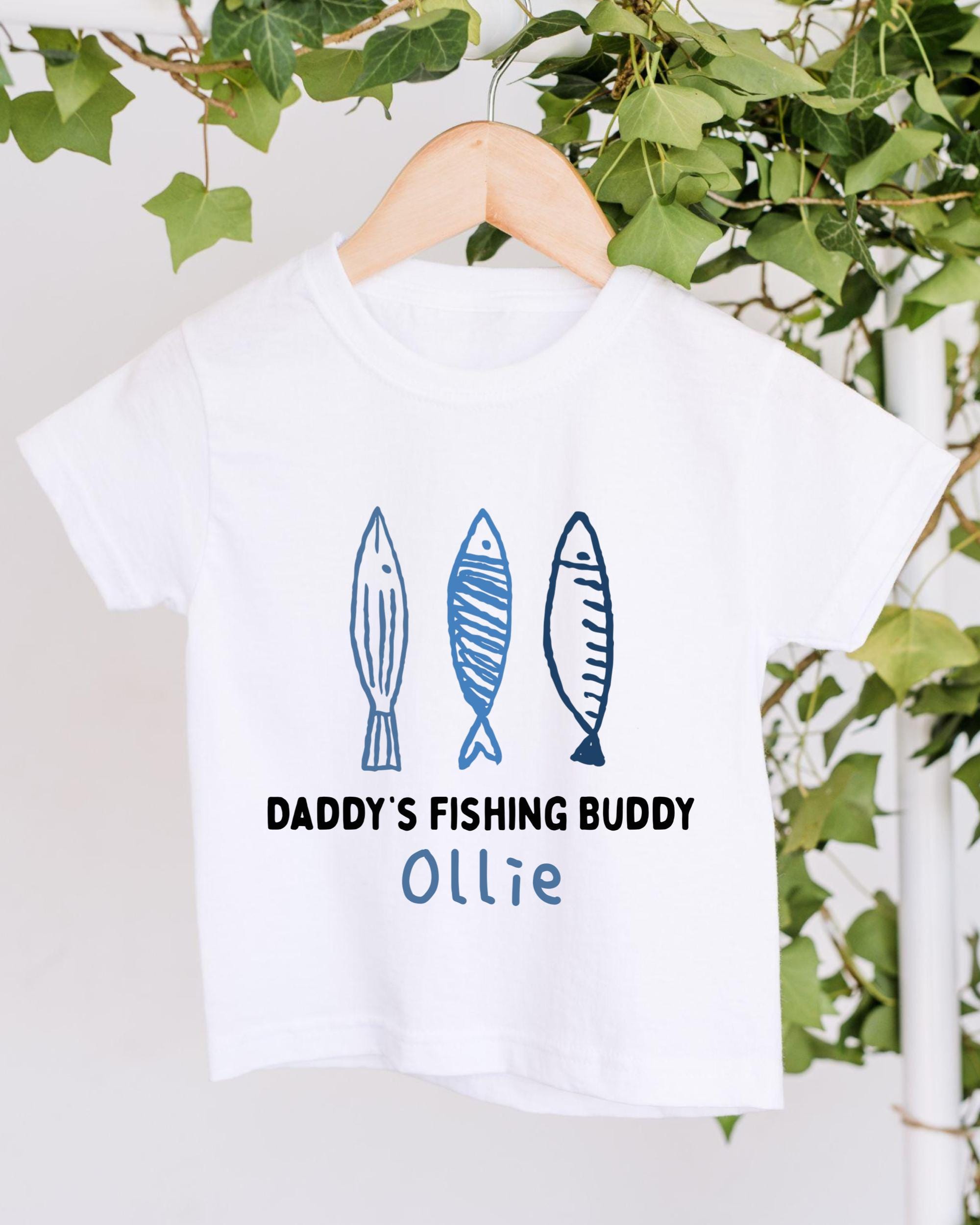 A white t-shirt with a graphic design featuring three fish and the text "DADDY'S FISHING BUDDY OLLIE" printed on it.