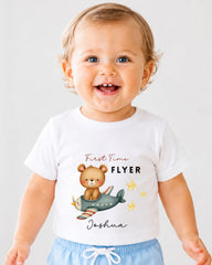 A smiling baby boy wearing a white t-shirt with a cartoon image of a bear on an airplane and the text "First Time Flyer" and "Joshua" printed on it.