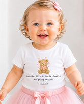 A smiling baby girl wearing a white t-shirt with a teddy bear graphic and text that says "Auntie Lily & Uncle Josh's are getting married! 20.10.2029".