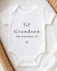 A white onesie with the text "First Grandson Due September 28" printed on it, placed on a white surface.