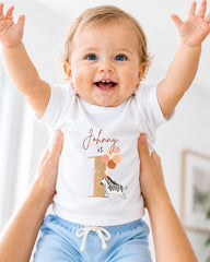 A smiling baby wearing a white t-shirt with a personalized number 1 and zebra design, being held up by someone's arms.