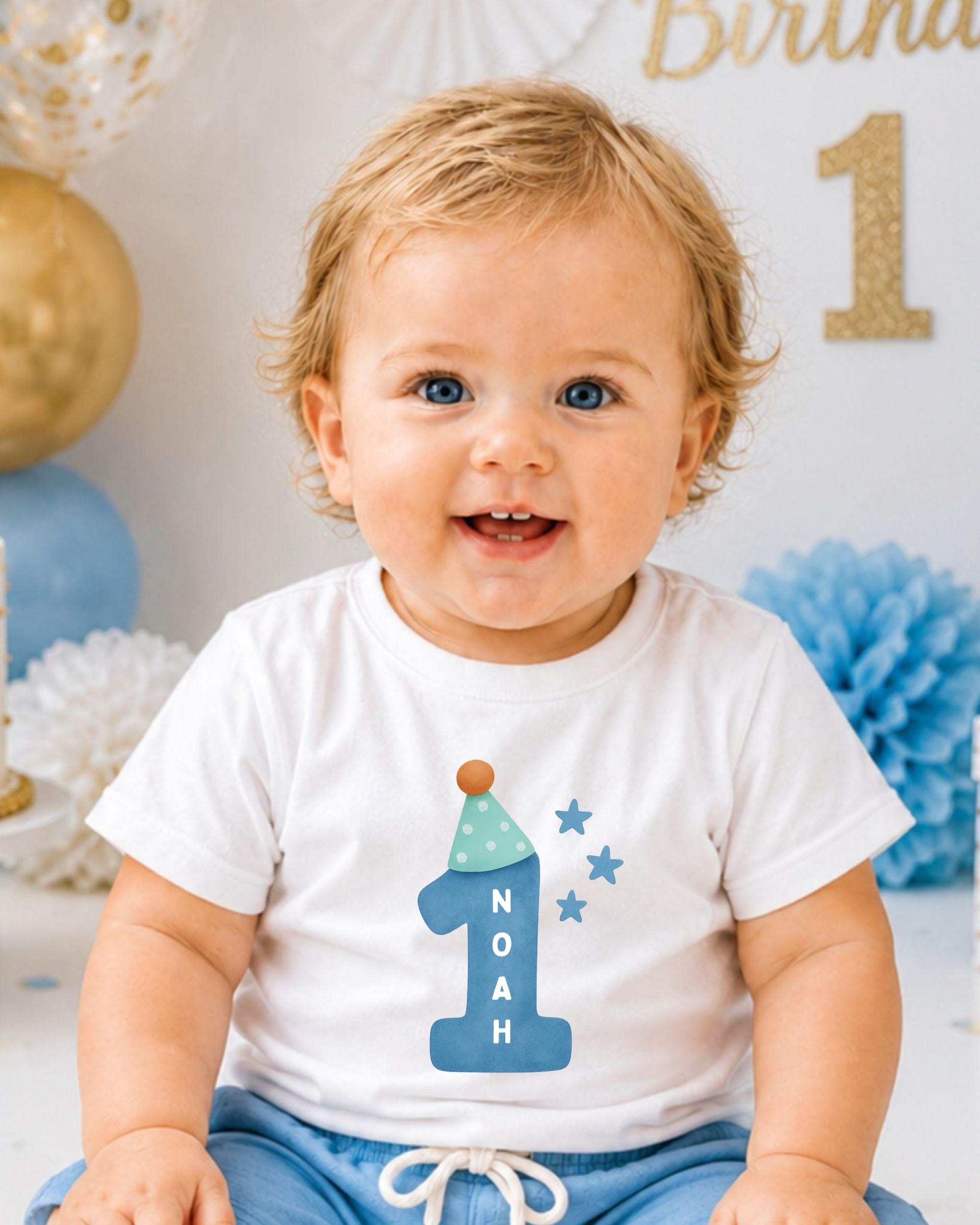 A smiling baby boy wearing a white t-shirt with a blue hat and stars on it, sitting in front of a birthday party backdrop.
