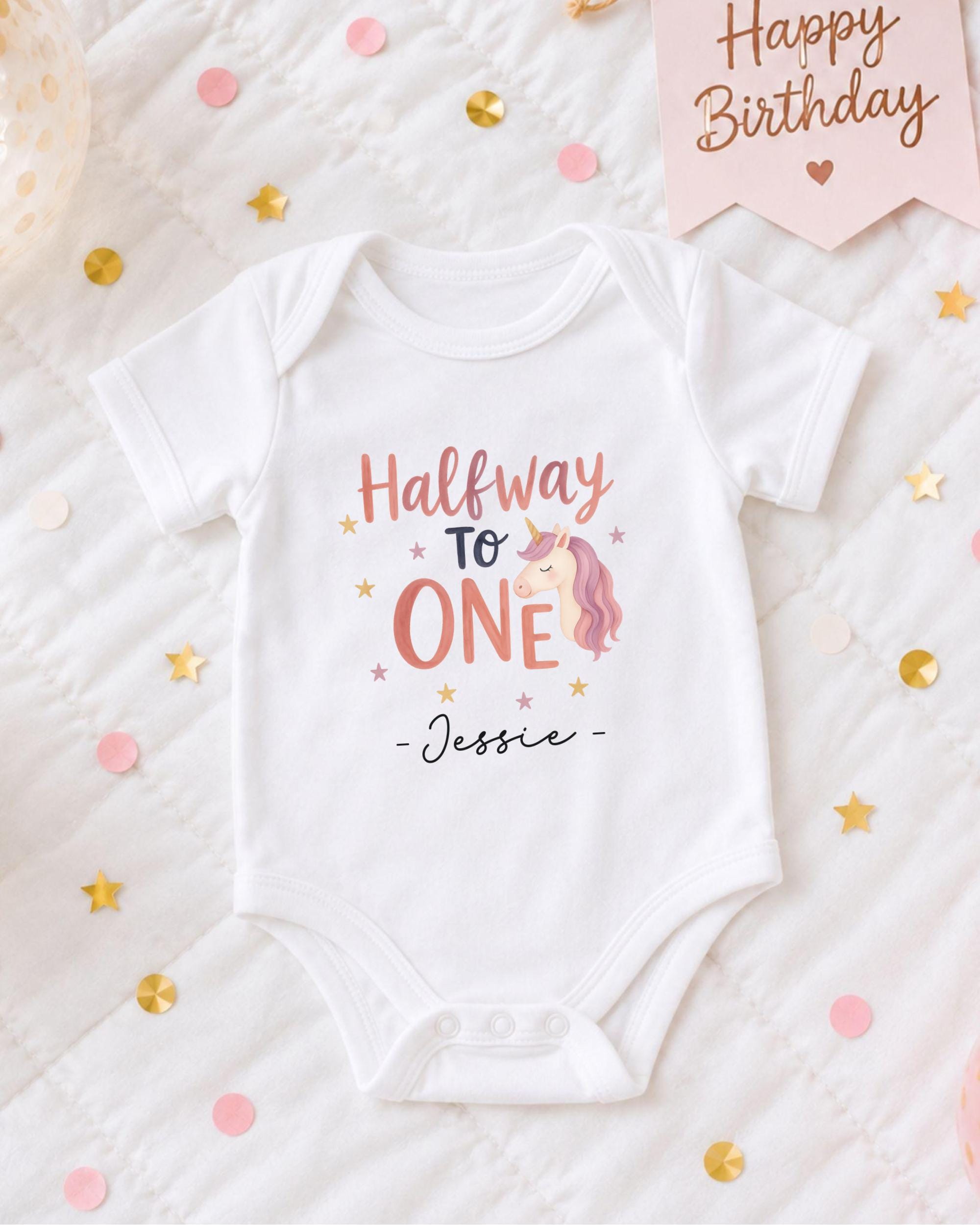 A white onesie with a unicorn design and the text "Halfway to One" printed on it, along with the name "JESSIE" below the unicorn image. The onesie is placed on a bed with confetti and a birthday card.
