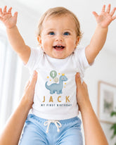 A smiling baby wearing a white shirt with a dinosaur and the text "Jack" and "My first birthday" printed on it, being held up by someone's arms.
