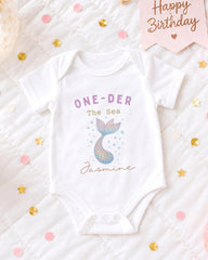 A white onesie with a mermaid design and the text "One-DER The Sea Jasmine" printed on it, placed on a bed with confetti and a "Happy Birthday" sign.