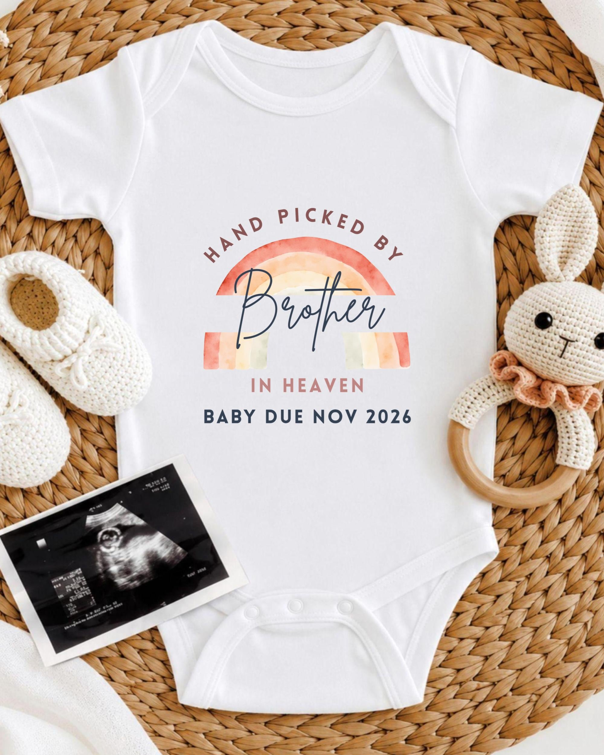 A white baby onesie with a rainbow graphic and text, along with a crocheted bunny toy and a baby's firstborn sibling announcement.
