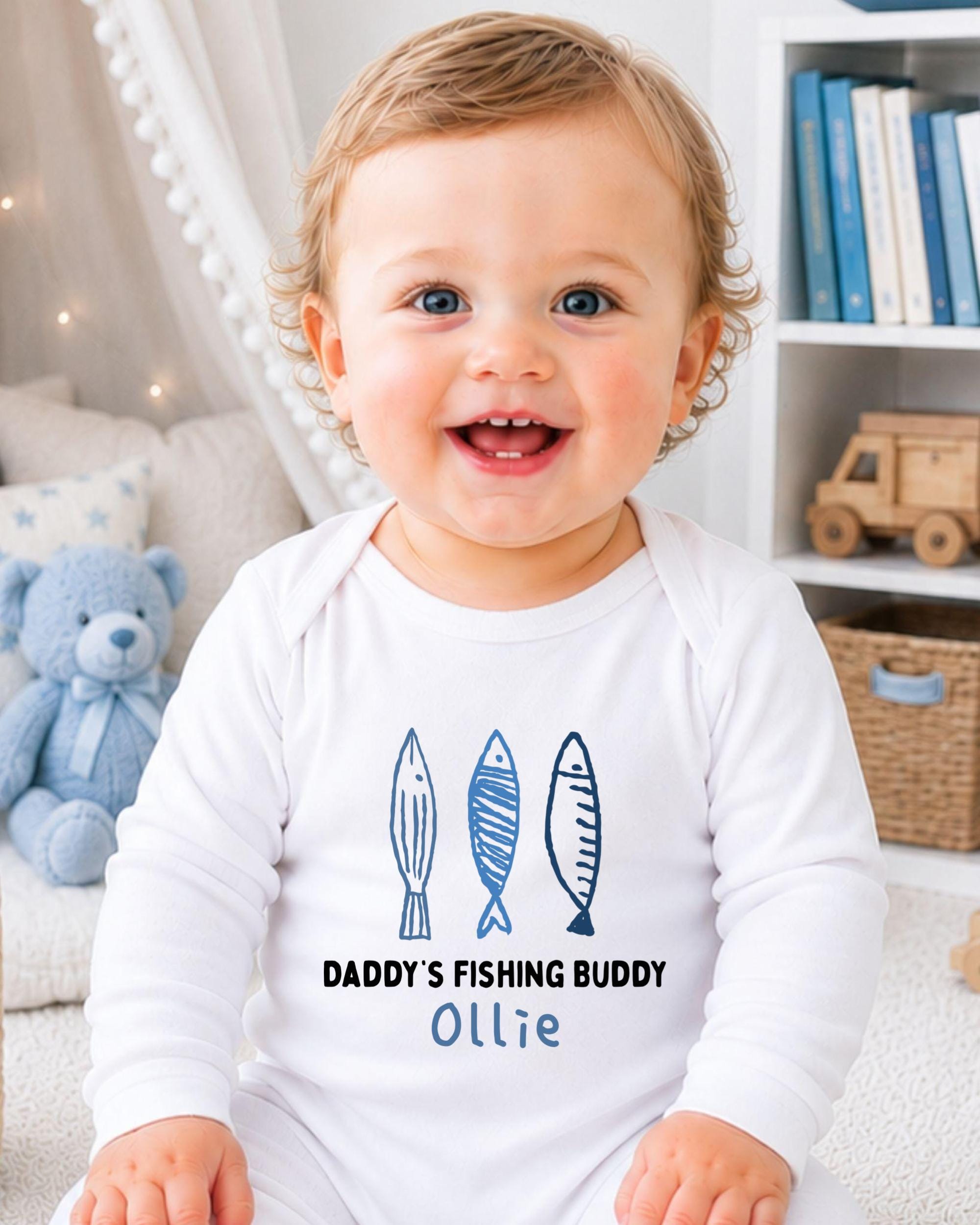 A smiling baby boy wearing a white onesie with a fishing theme and the text "DADDY'S FISHING BUDDY OLLIE" printed on it.
