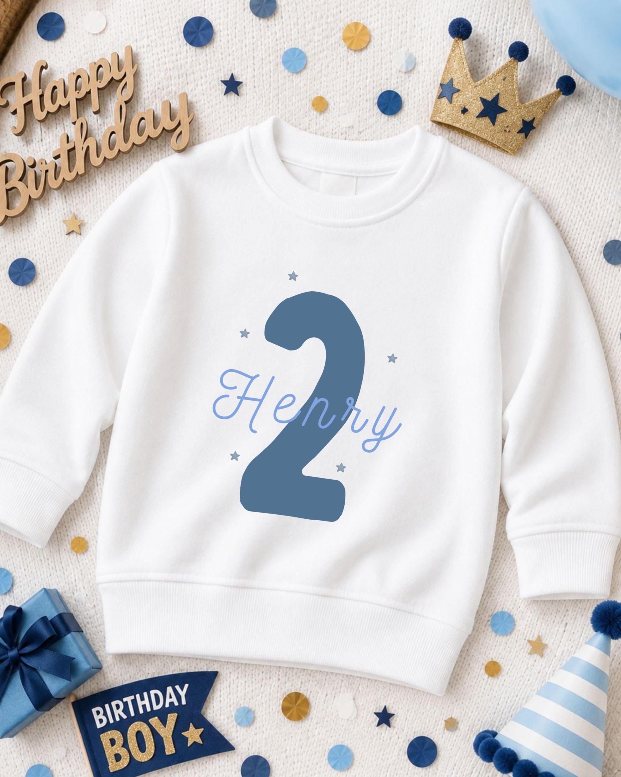 A white sweatshirt with the number "2" and the name "Henry" printed on it, surrounded by birthday decorations such as a crown, confetti, and a birthday hat.