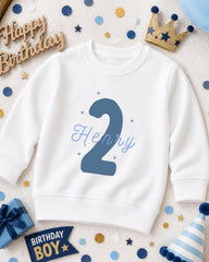 A white sweatshirt with the number "2" and the name "Henry" printed on it, surrounded by birthday decorations such as a crown, confetti, and a birthday hat.