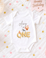 A white onesie with the text "amy is one" printed on it, surrounded by confetti and a birthday card.