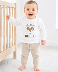 A smiling baby boy wearing a white sweatshirt with a graphic of a ram and the text " Justin's 1st Rodeo" standing in a nursery.