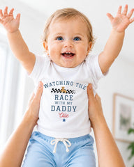 A smiling baby wearing a white shirt with the text "WATCHING THE RACE WITH MY DADDY" is being held up by someone's arms.