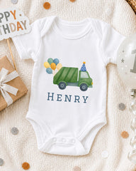 A white onesie with a green truck and balloons on it, along with a "Happy Birthday" sign and a birthday balloon.