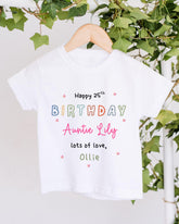A white t-shirt with colorful text and hearts hanging on a wooden hanger, with greenery in the background.