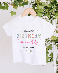 A white t-shirt with colorful text and hearts hanging on a wooden hanger, with greenery in the background.