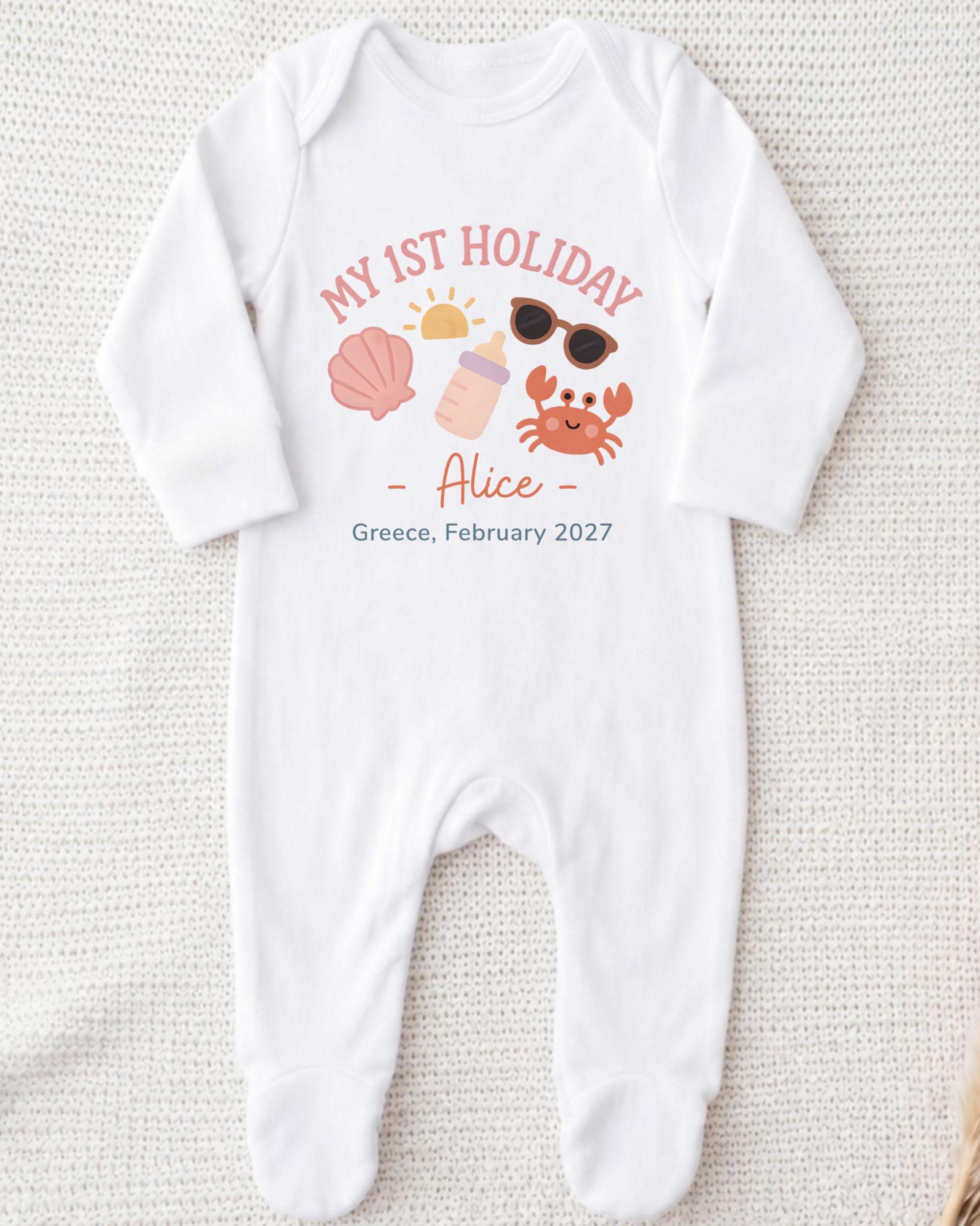 A white onesie with a personalized design on it, featuring a sun, sunglasses, a shell, and a crab, along with the text "My 1st Holiday - Alice - Greece, February 2027".