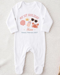 A white onesie with a personalized design on it, featuring a sun, sunglasses, a shell, and a crab, along with the text "My 1st Holiday - Alice - Greece, February 2027".