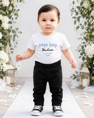 A young child wearing a white t-shirt with the text "Page Boy Hudson" and black pants stands in front of a floral backdrop.