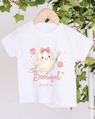 A white t-shirt with a cartoon ghost character and the text "Little Bootful Annie" printed on it, hanging on a wooden hanger against a backdrop of greenery.